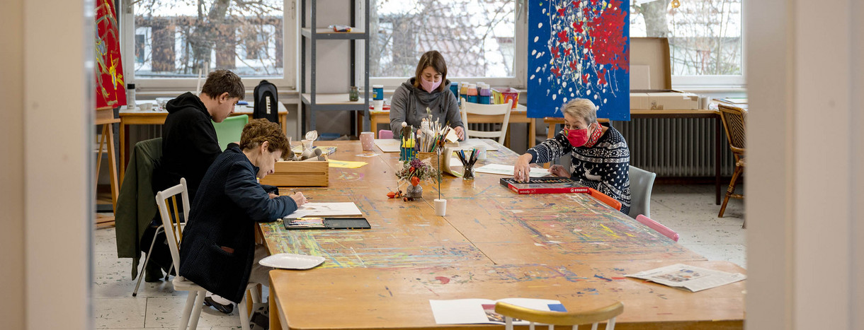 An einem Tisch großen Tisch in einer Kunstwerkstatt arbeiten Künstler an Bildern.