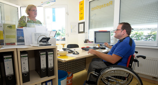 Ein Mann im Rollstuhl arbeitet an der Rezeption eines Campingplatzes. (Foto: © dpa)']