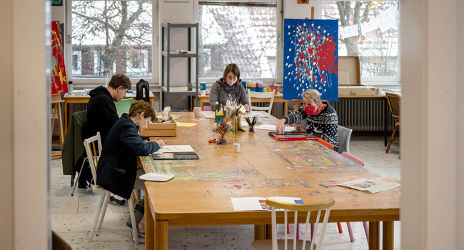 An einem Tisch großen Tisch in einer Kunstwerkstatt arbeiten Künstler an Bildern.']